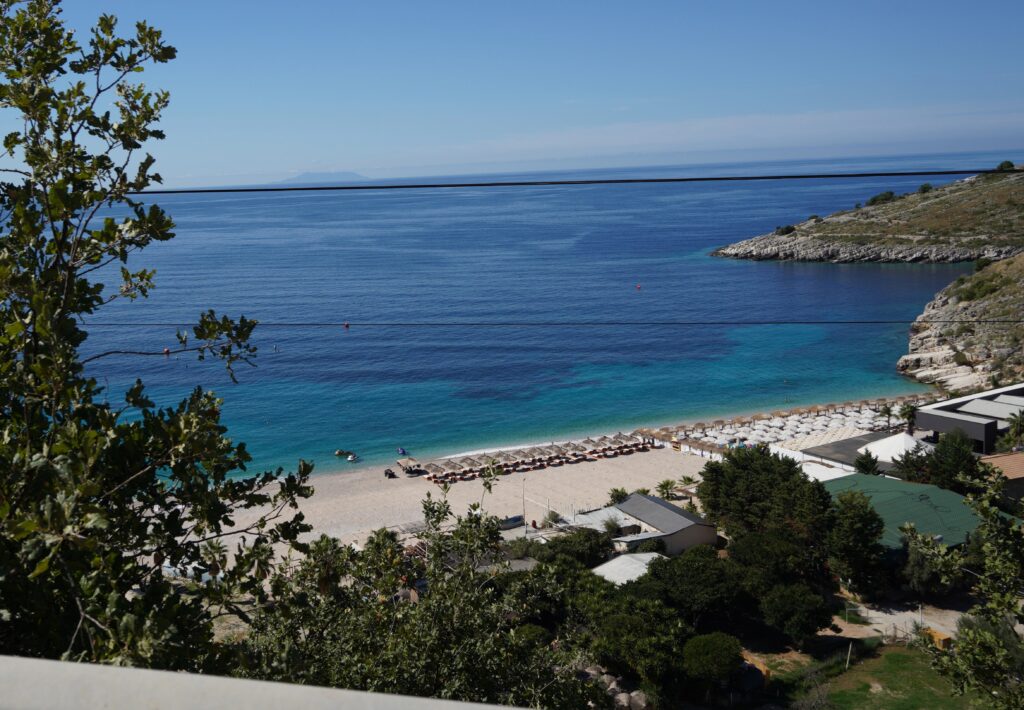 Secluded beach with umbrellas and turquoise water near Himarë, Albanian Riviera, seen from the SH8 coastal road.