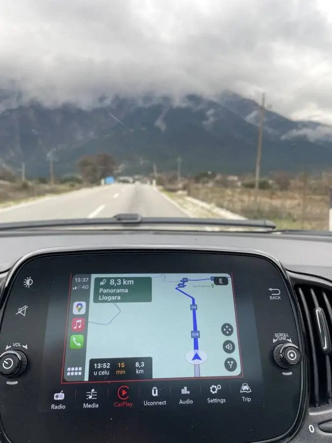 Navigation screen showing route to Llogara Pass (SH8) in Albania, with dramatic mountain backdrop — a scenic but demanding drive often requiring full rental insurance coverage.
